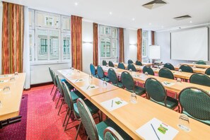 Un salón de conferencias con varias mesas y sillas, ventanas amplias con cortinas y una pizarra.