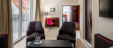 A hotel room with a large window, two armchairs, a coffee table, a desk, and a TV.