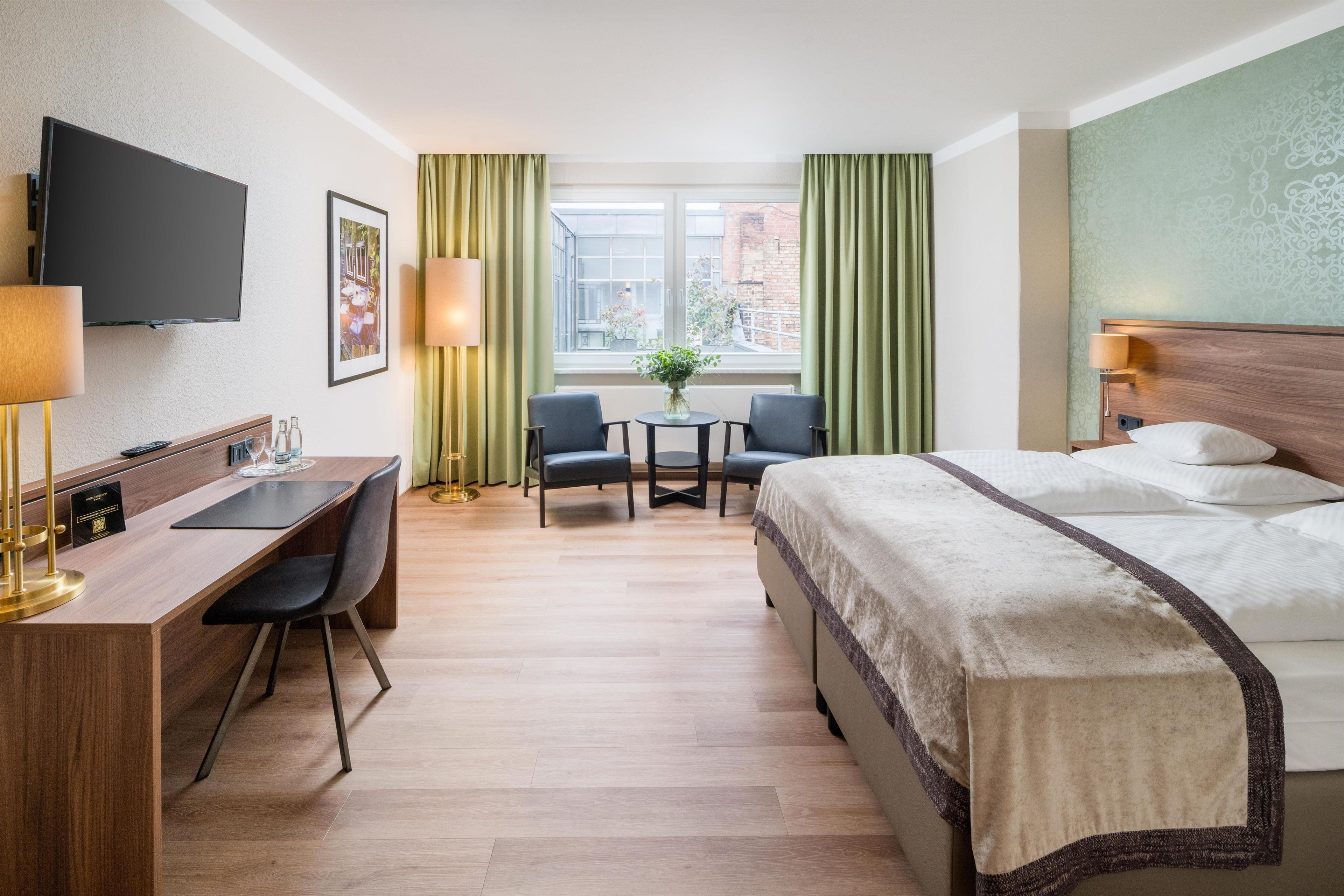 Une chambre d’hôtel avec un lit, un bureau, des chaises, une télévision et une fenêtre avec des rideaux.