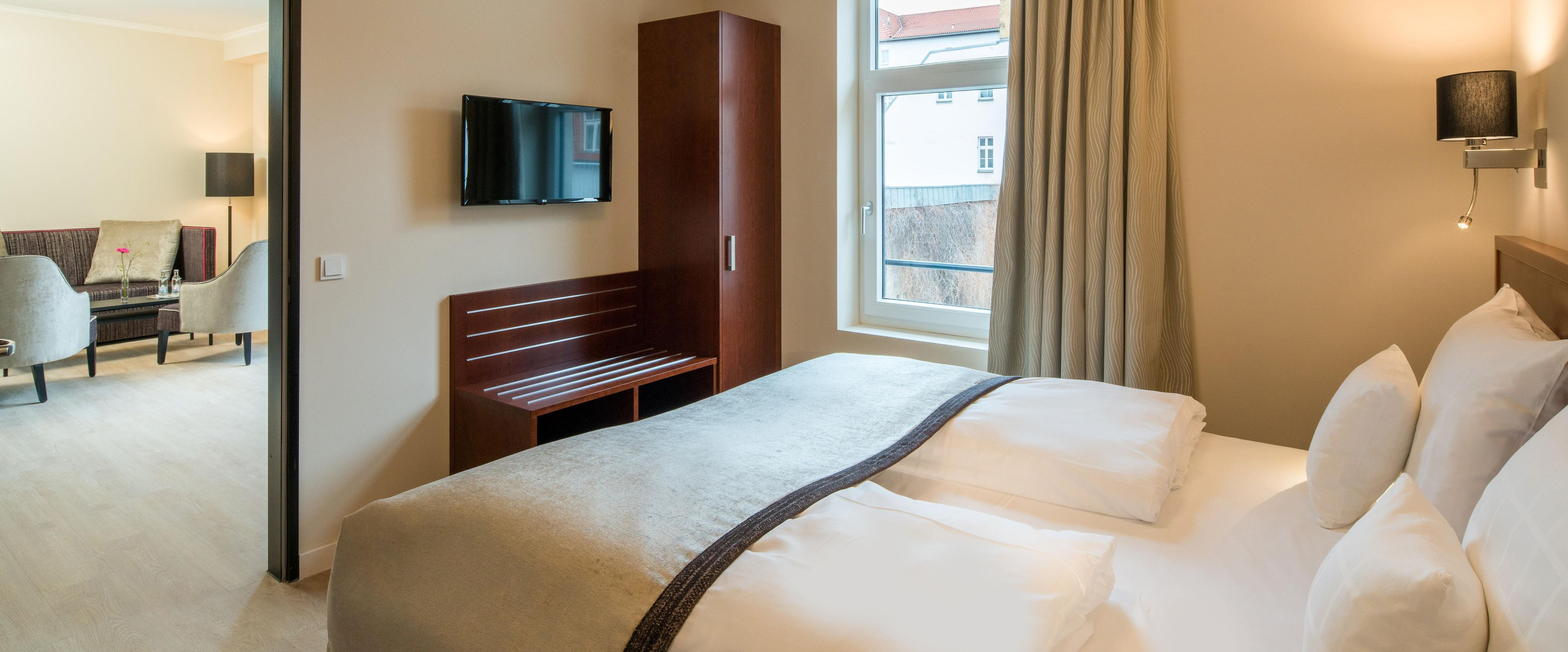 Habitación de hotel con cama, televisor montado en la pared, banca de madera y ventana con cortinas.