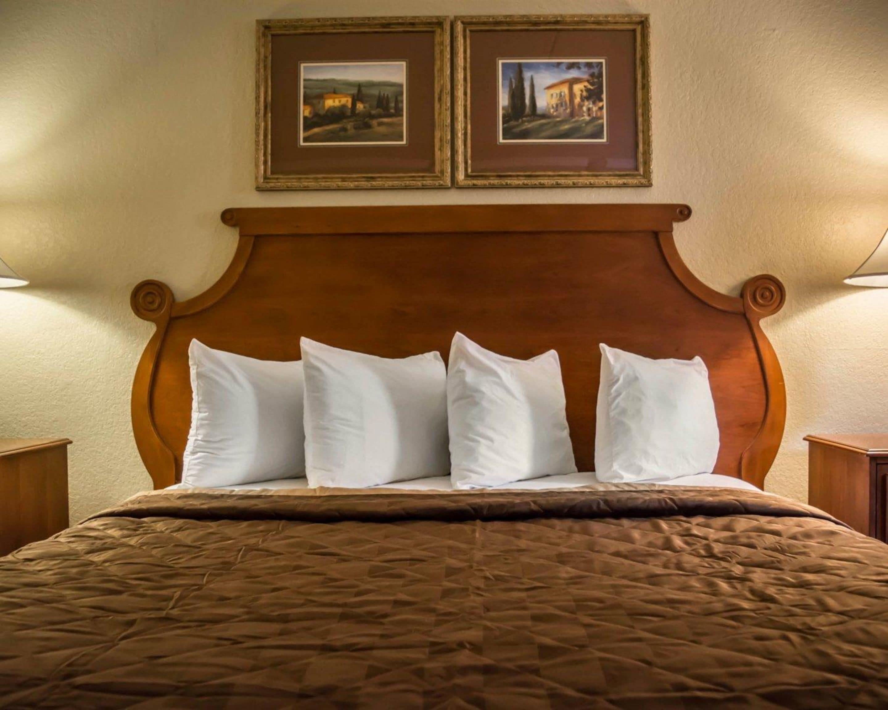 A hotel room with a wooden headboard, four white pillows, a brown quilted bedspread, and two framed pictures on the wall.