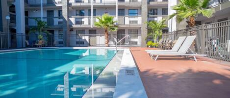A swimming pool with lounge chairs and palm trees, surrounded by a multi-story building with balconies.