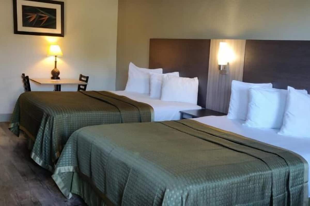 A hotel room with two beds, each with white linens and green bedspreads. There are two bedside tables with lamps, a chair, and a framed picture on the wall.