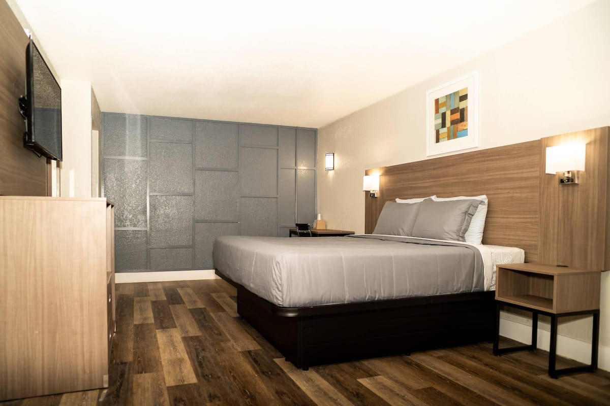 A modern hotel room with a large bed, a flat-screen TV mounted on the wall, and a small bedside table with a lamp.