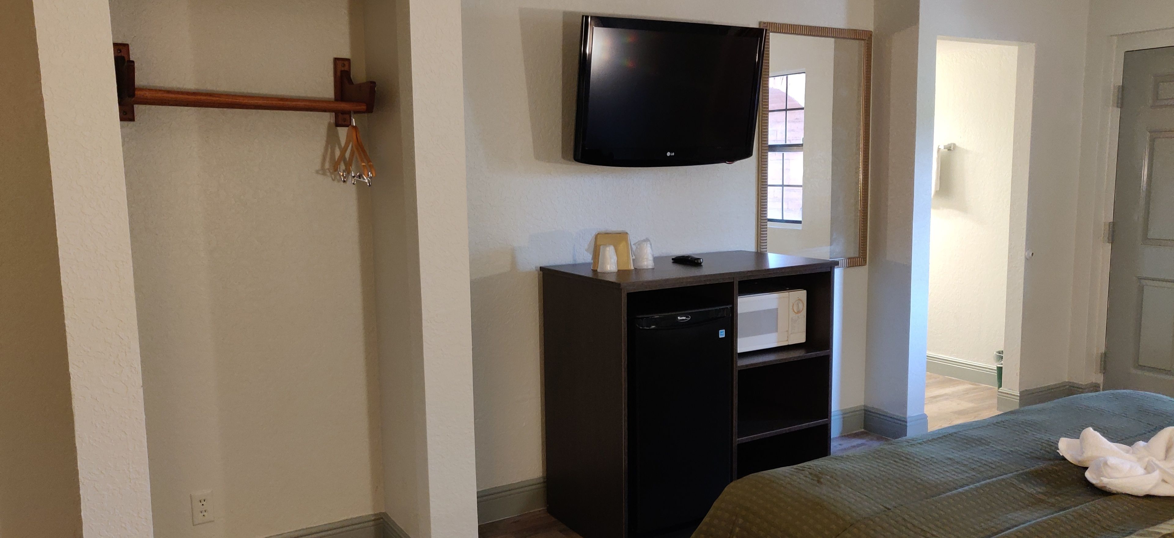 A hotel room with a TV mounted on the wall, a small desk with a refrigerator, and a bed with a blanket.