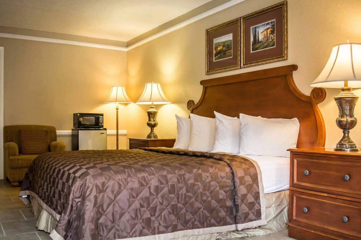 A hotel room with a bed, two bedside lamps, a TV, a chair, and a wooden nightstand with drawers.