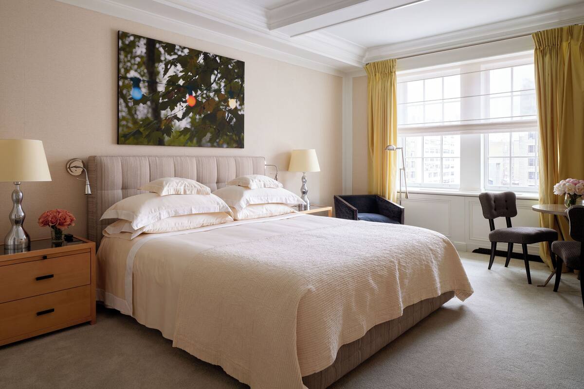 A hotel room with a large bed, two bedside tables with lamps, a chair, a small table with a vase, and a large window with curtains.