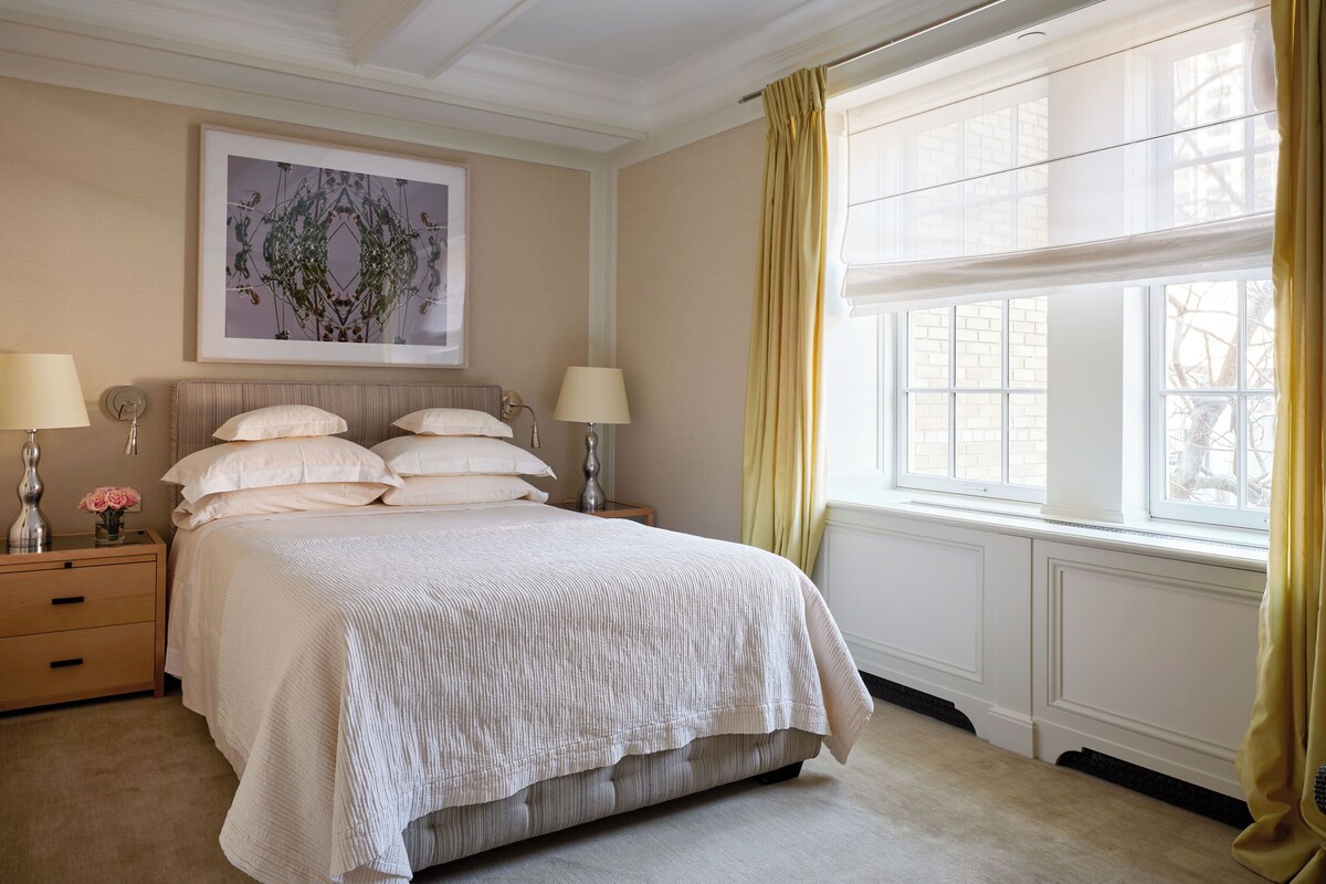 Un dormitorio ordenado con una cama grande, dos mesitas de noche con lámparas, una obra de arte enmarcada sobre la cama y una ventana con cortinas.