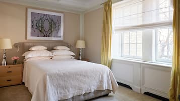 Un dormitorio ordenado con una cama grande, dos mesitas de noche con lámparas, una obra de arte enmarcada sobre la cama y una ventana con cortinas.