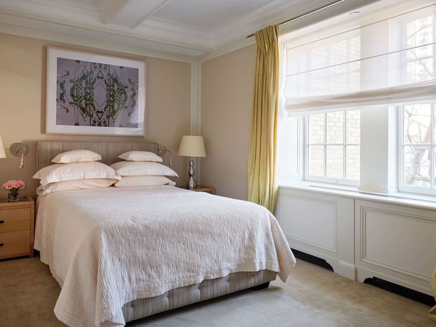 Un dormitorio ordenado con una cama grande, dos mesitas de noche con lámparas, una obra de arte enmarcada sobre la cama y una ventana con cortinas.