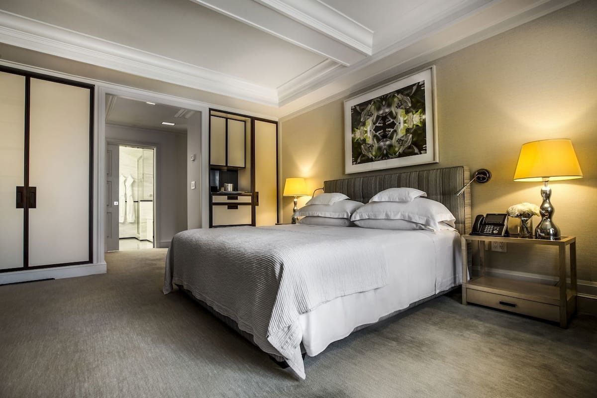 A hotel room with a large bed, bedside lamps, a nightstand, a framed artwork on the wall, and a sliding door leading to another room.