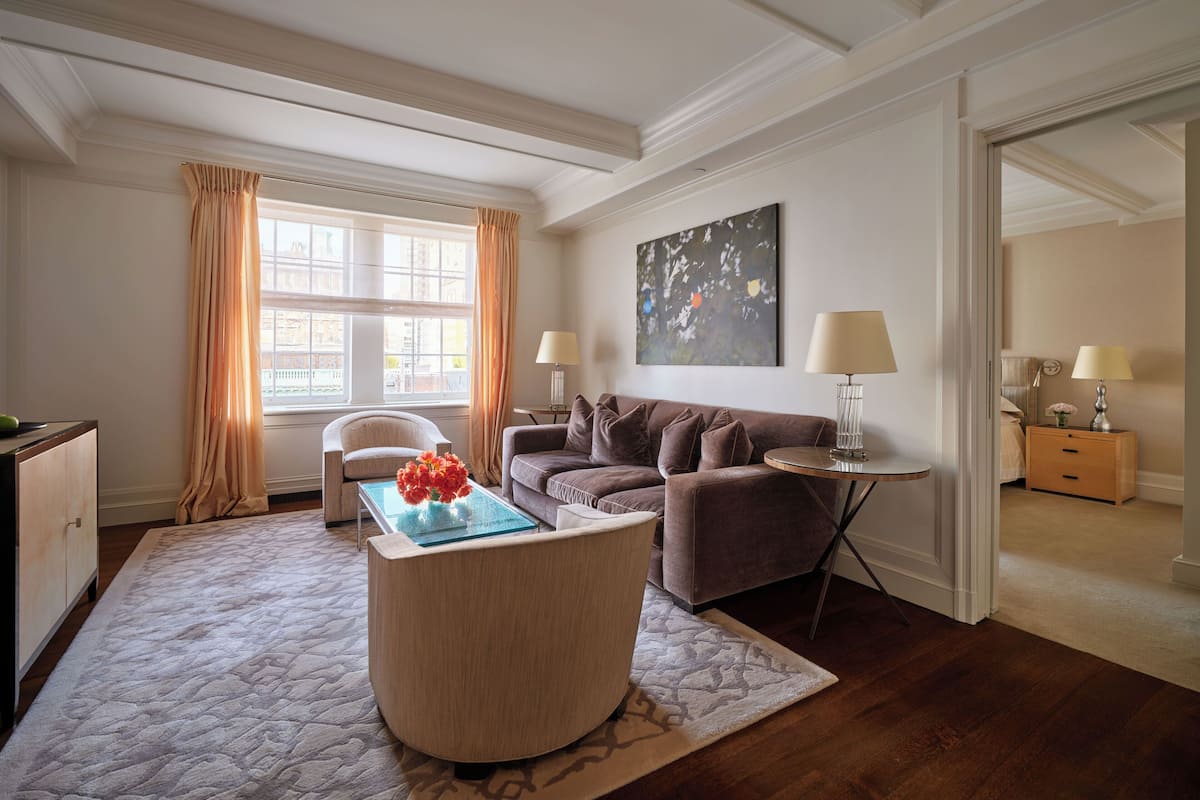 A living room with a sofa, coffee table, and lamps. There is a window with curtains and a painting on the wall.