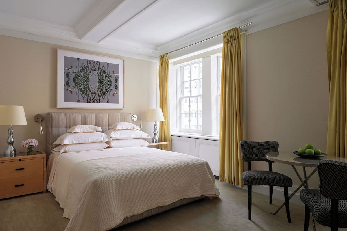 A hotel room with a bed, bedside tables, a desk with chairs, a window with curtains, and a framed artwork on the wall.