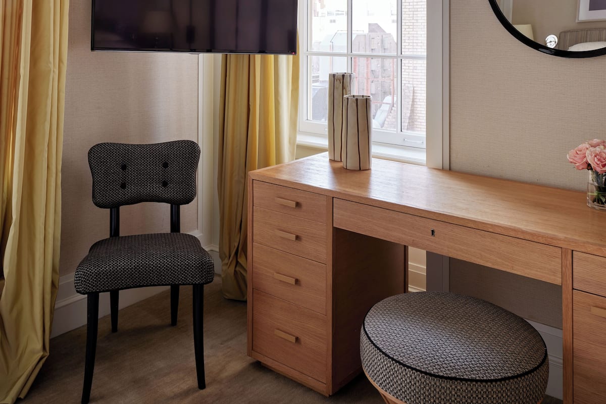 A room with a wooden desk, a chair, a round ottoman, a TV mounted on the wall, and a window with yellow curtains.