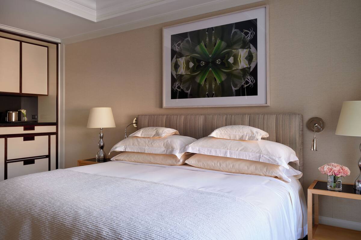 A neatly made bed with white linens, a large abstract painting above it, and a small table with a vase of flowers beside the bed.