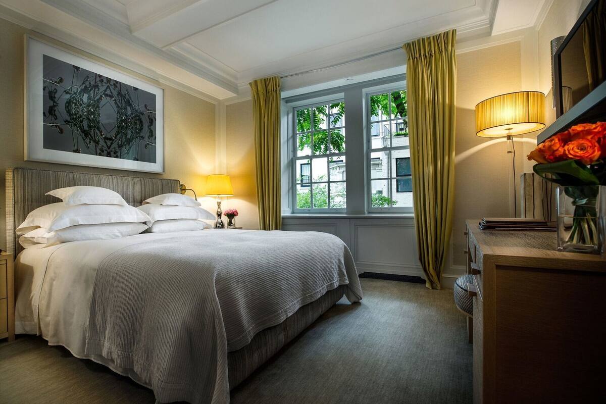 A hotel room with a bed, a nightstand, a lamp, a vase with flowers, a painting on the wall, and a window with curtains.