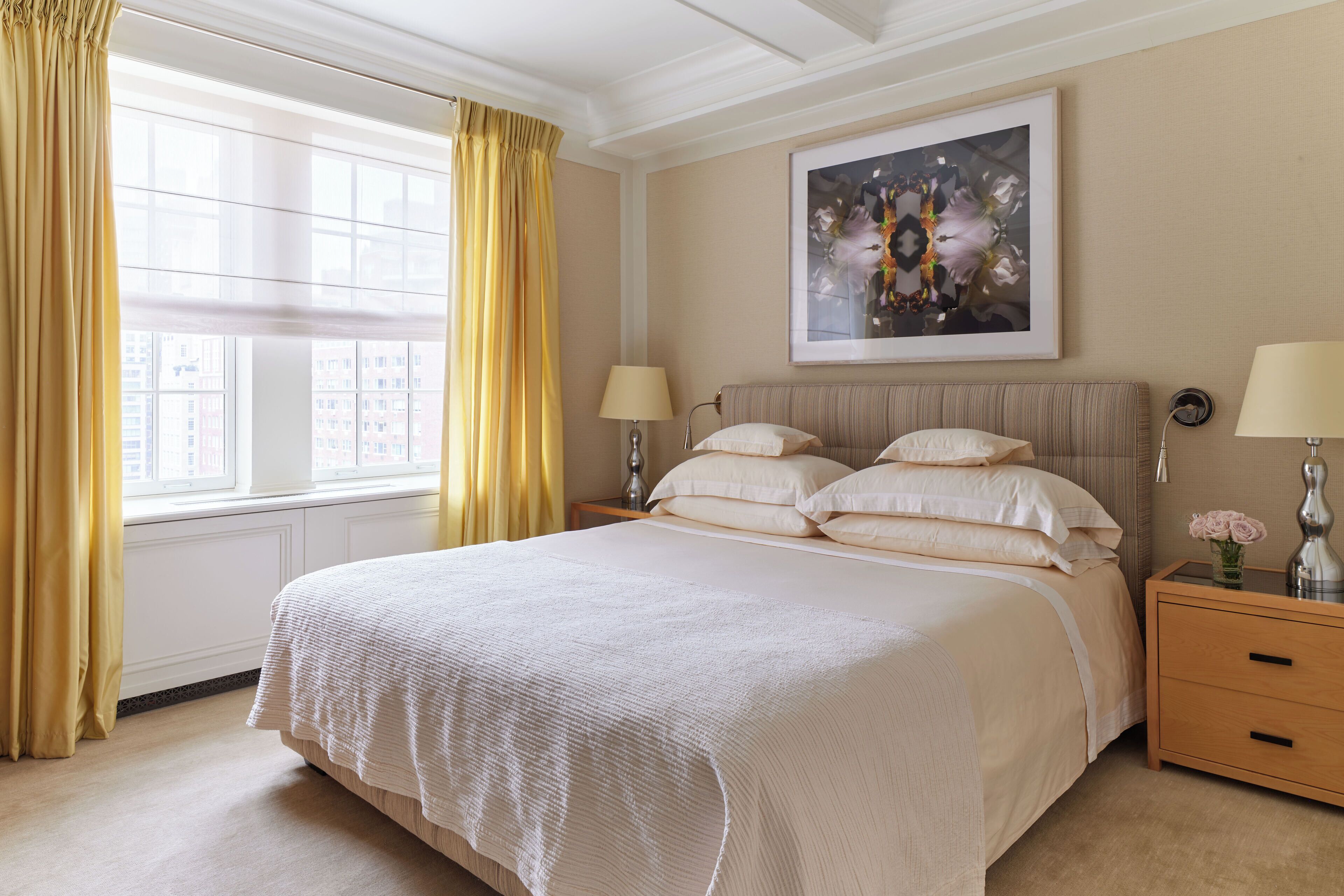 Una cama grande con cubrecama blanco, dos mesitas de noche con lámparas, una obra de arte enmarcada sobre la cama y una ventana con cortinas amarillas.