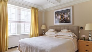 A large bed with a white bedspread, two bedside tables with lamps, a framed artwork above the bed, and a window with yellow curtains.