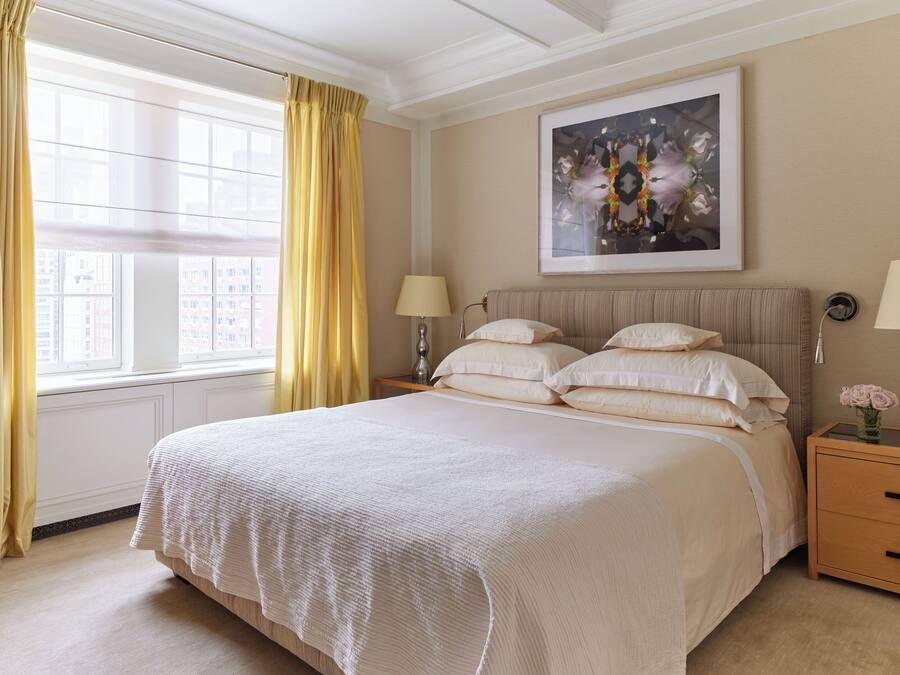 Een groot bed met een witte spreidek, twee nachtkastjes met lampen, een kunstwerk in een lijst boven het bed, en een raam met gele gordijnen.
