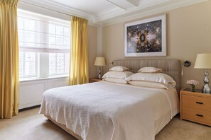 A large bed with a white bedspread, two bedside tables with lamps, a framed artwork above the bed, and a window with yellow curtains.