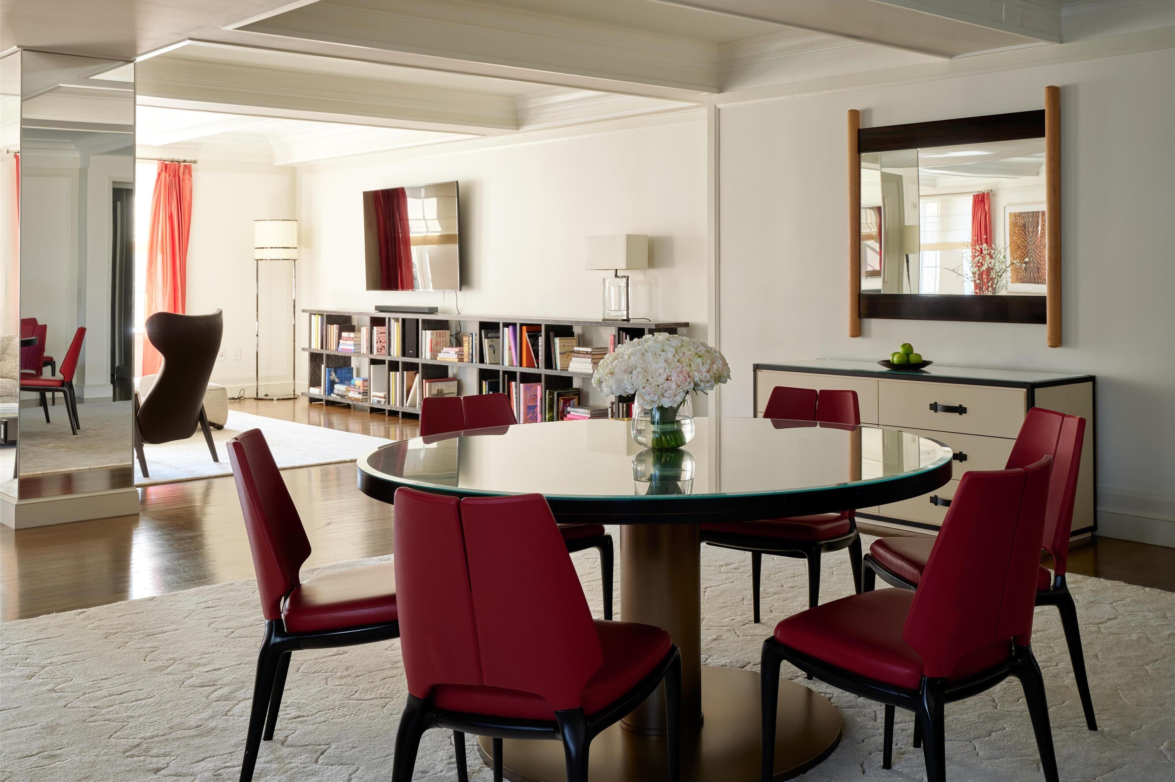 A modern dining area with a round glass table, red upholstered chairs, a vase of flowers, and a bookshelf in the background.