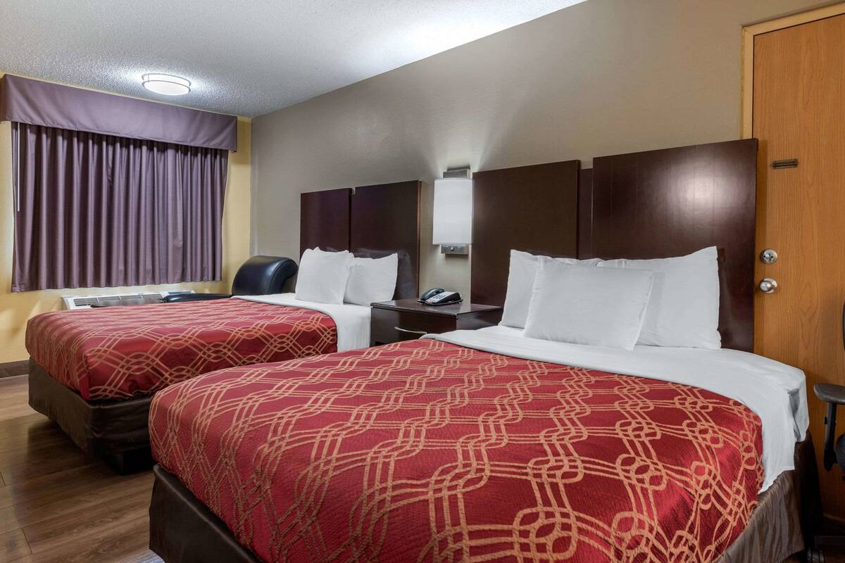 Quarto de hotel com duas camas, uma escrivaninha, uma cadeira e uma janela grande com cortinas.