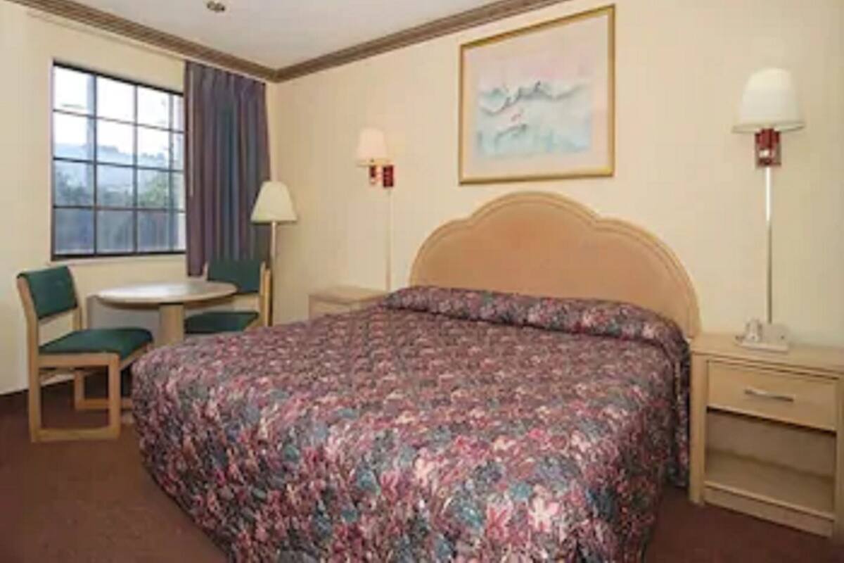Quarto de hotel com uma cama grande, uma escrivaninha, uma cadeira, uma janela com cortinas e um quadro na parede.