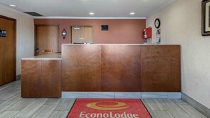 Et receptionsområde med en træreception, et rødt tæppe med EconoLodge logo, et ur og en brandslukker på væggen.