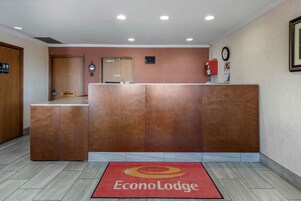Un área de recepción con un mostrador de madera, una alfombra roja con el logotipo de EconoLodge, un reloj y un extintor en la pared.