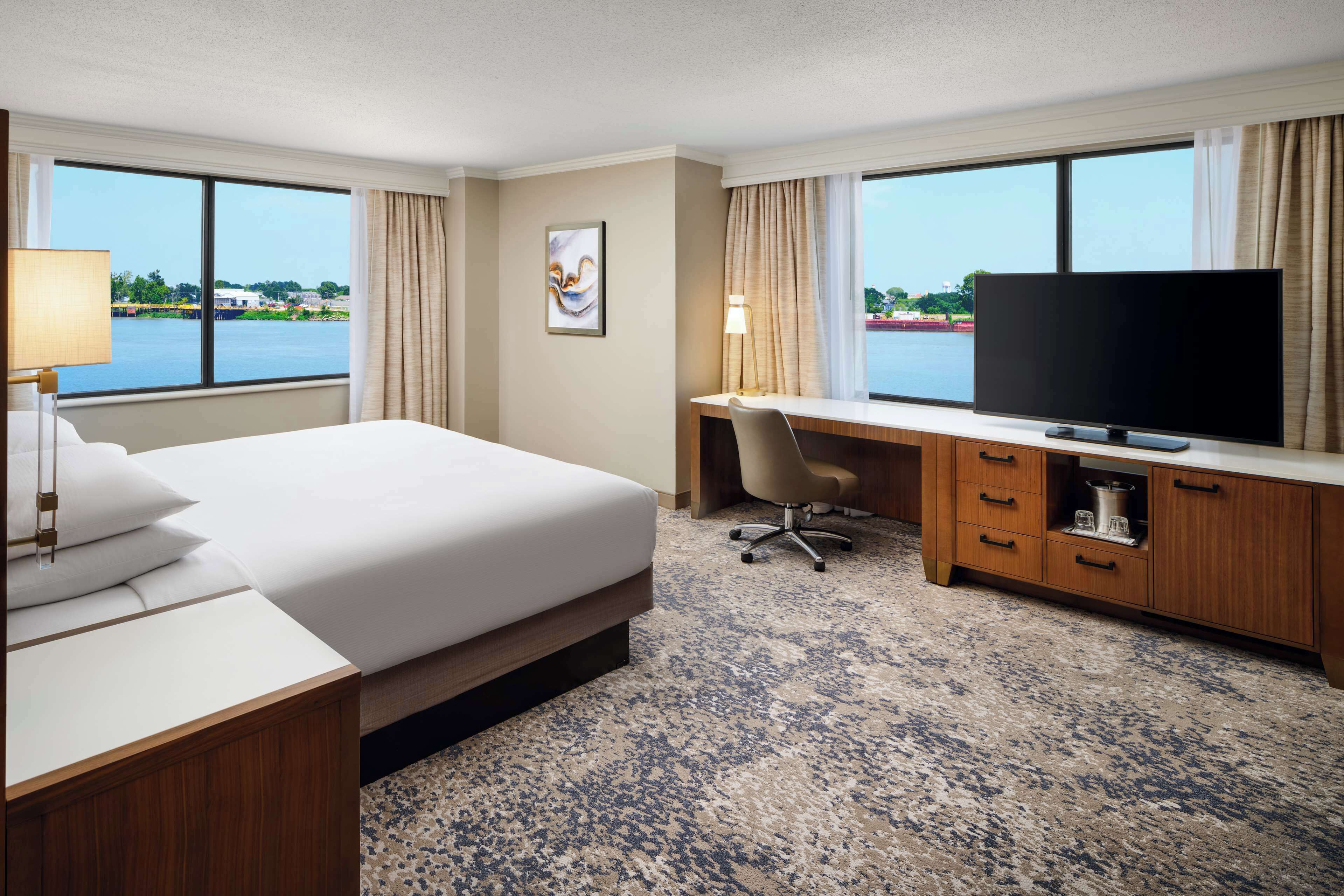 A hotel room with a bed, desk, television, and a view of a body of water.