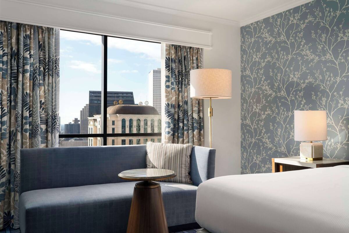 Habitación de hotel con un ventanal grande, un sofá, una mesa redonda, una lámpara y una cama.