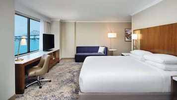 A hotel room with a bed, desk, chair, TV, and a view of a bridge.