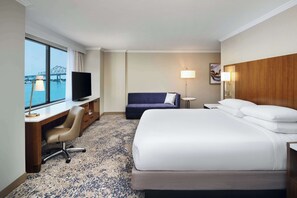 A hotel room with a bed, desk, chair, TV, and a view of a bridge.