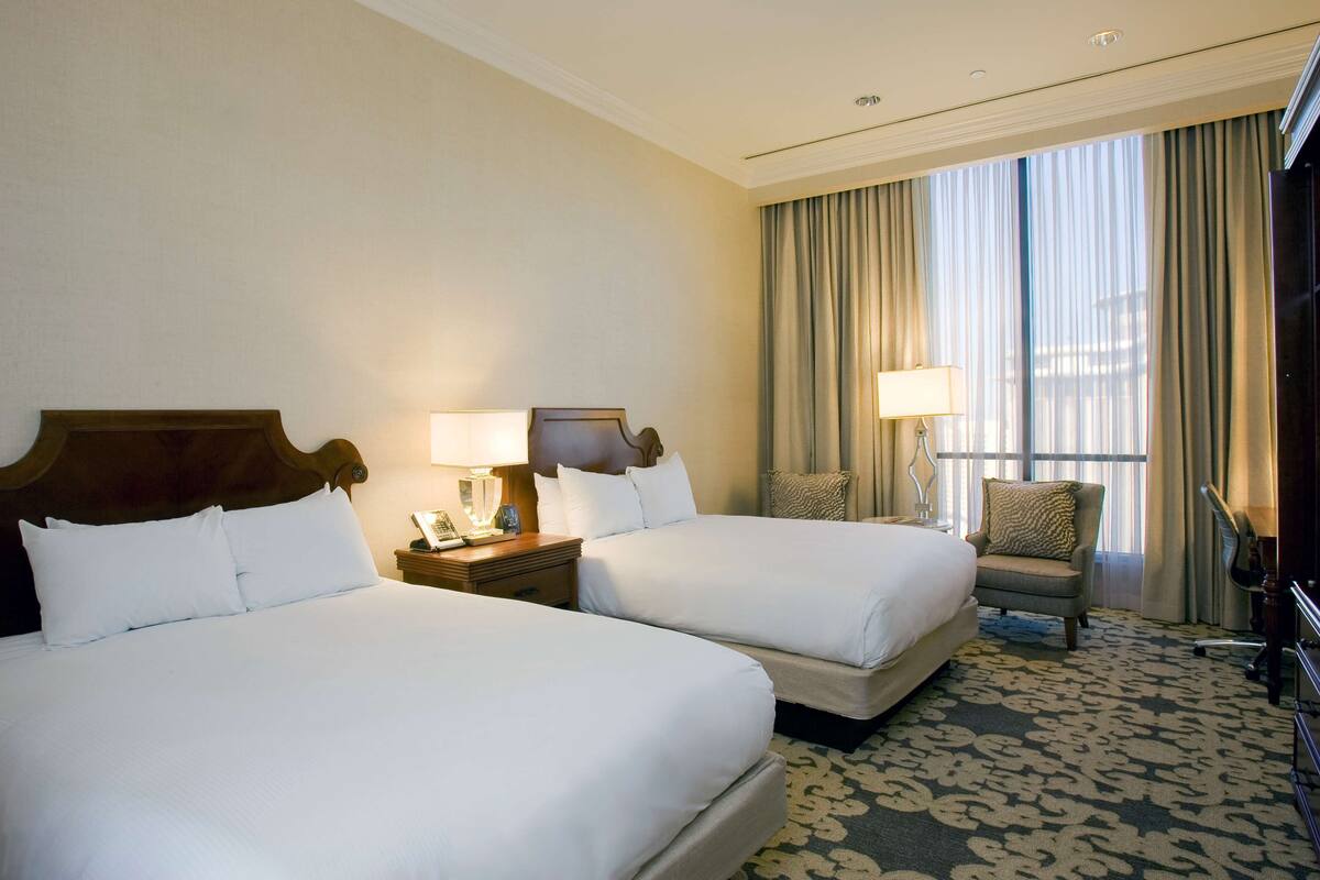 Habitación de hotel con dos camas, una silla, un escritorio y un ventanal grande con cortinas.