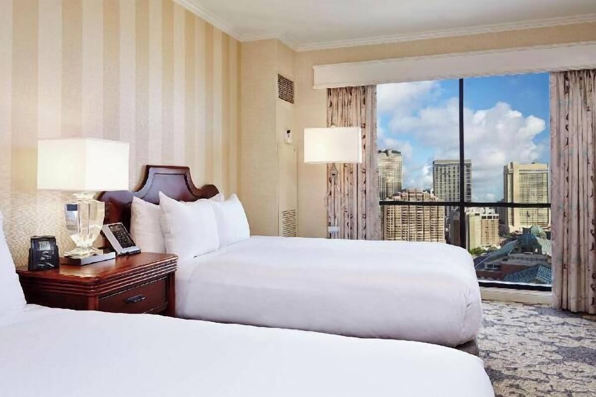 Habitación de hotel con dos camas, una mesita de noche con lámpara, teléfono y vistas a un paisaje urbano a través de amplias ventanas.