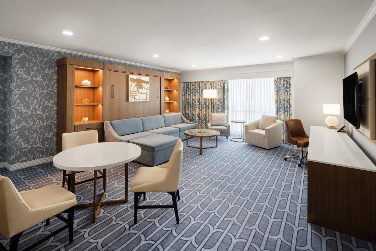 Una habitación de hotel moderna con sofá, sillones, una mesa de comedor, televisor y minibar.