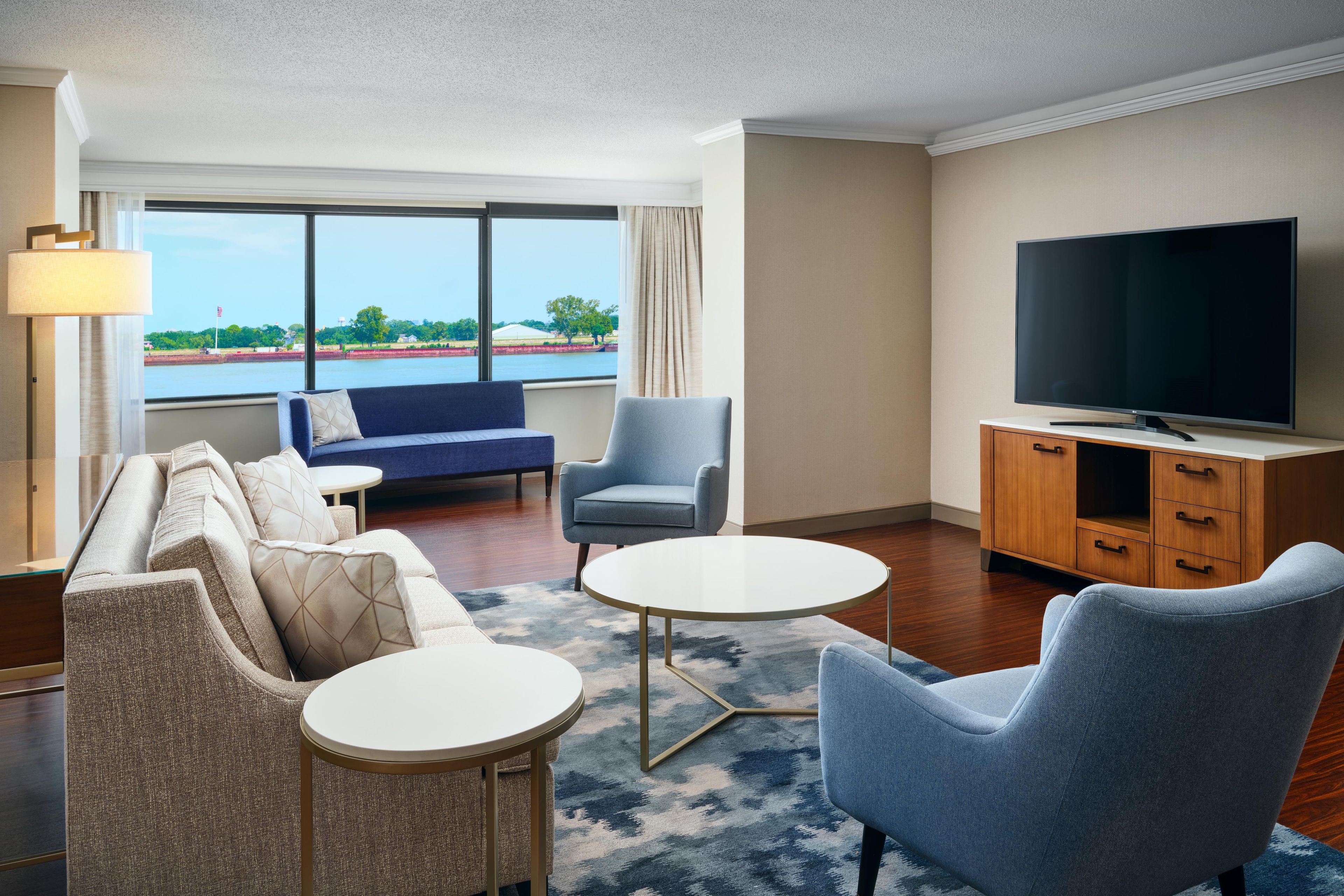 Hotelkamer met een zithoek, een fauteuil, een klein rond tafeltje, een televisie en uitzicht op een wateroppervlak door grote ramen.