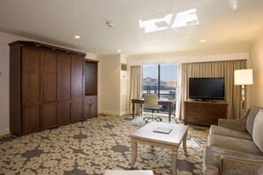 A hotel room with a large wardrobe, a flat-screen TV, a sofa, a coffee table, and a view of a bridge through the window.