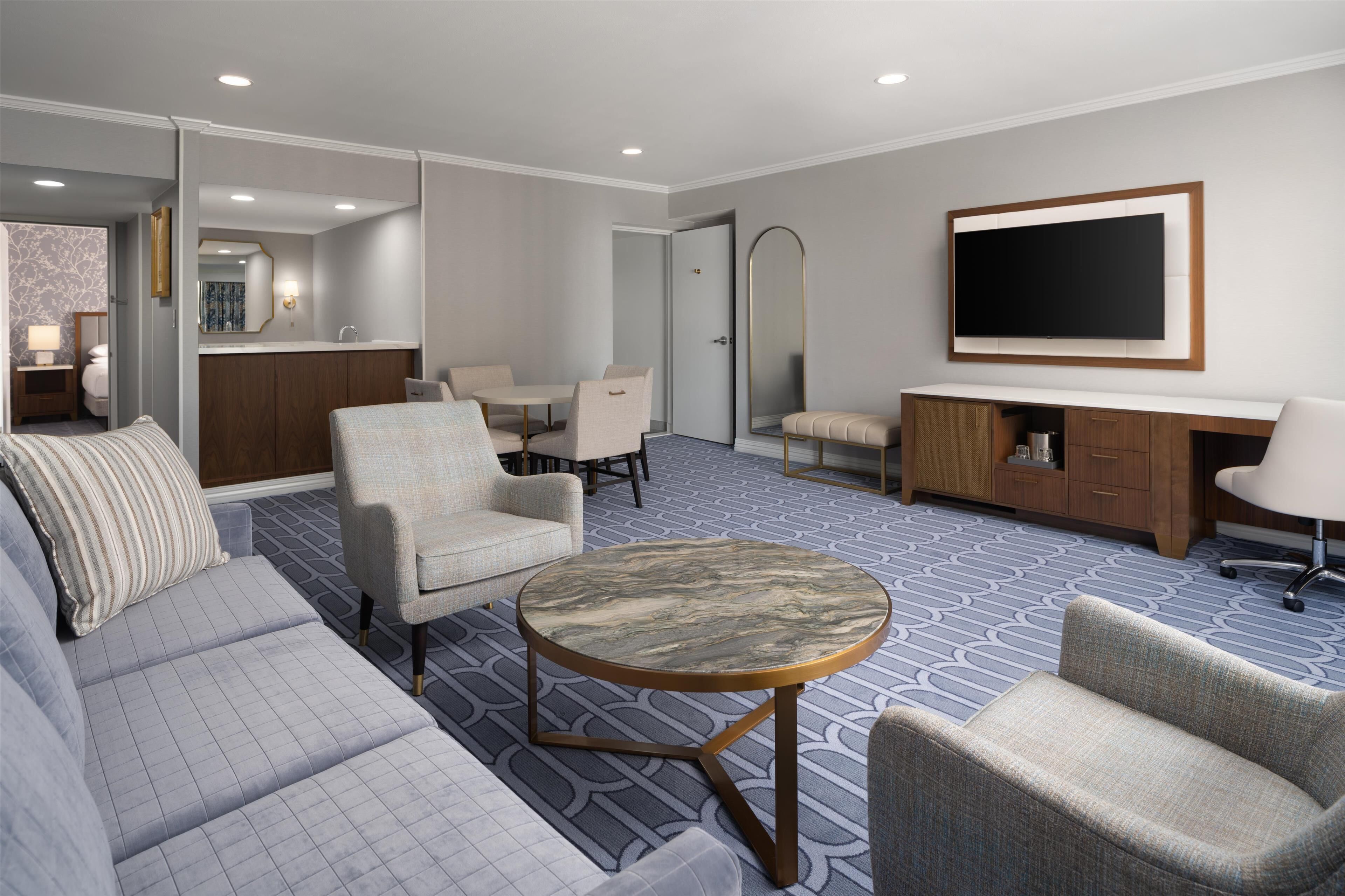 A hotel room with a sofa, armchairs, a round table, a TV, a mirror, a minibar, and a bathroom visible through an open door.