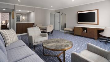 A hotel room with a sofa, armchairs, a round table, a TV, a mirror, a minibar, and a bathroom visible through an open door.