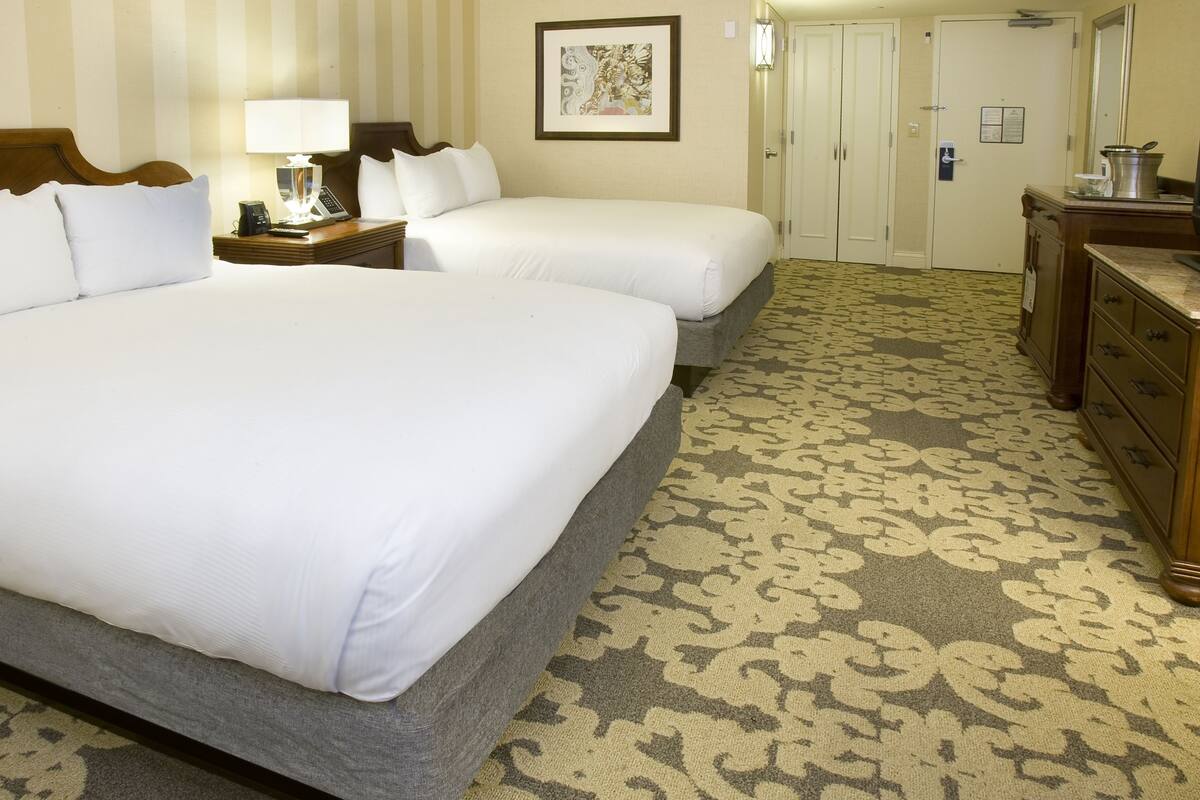 Una habitación de hotel con dos camas, una mesita de noche con lámpara, un aparador y una alfombra estampada.