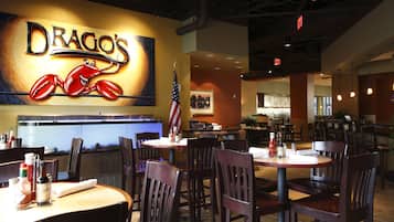 A restaurant interior with tables and chairs, a large sign reading "DRAGO'S" with a crab mascot, an American flag, and a counter area.