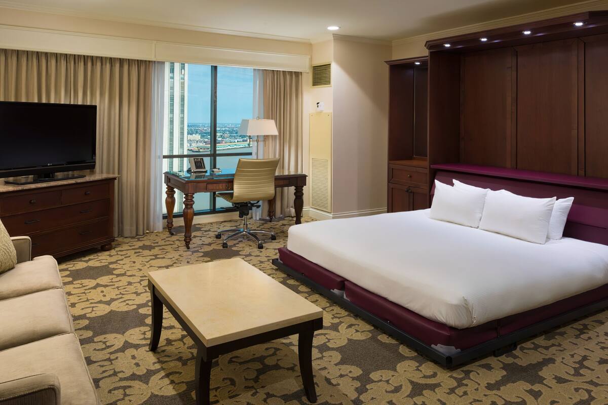 Habitación de hotel con una cama grande, un escritorio con silla, un televisor y vista a la ciudad desde la ventana.