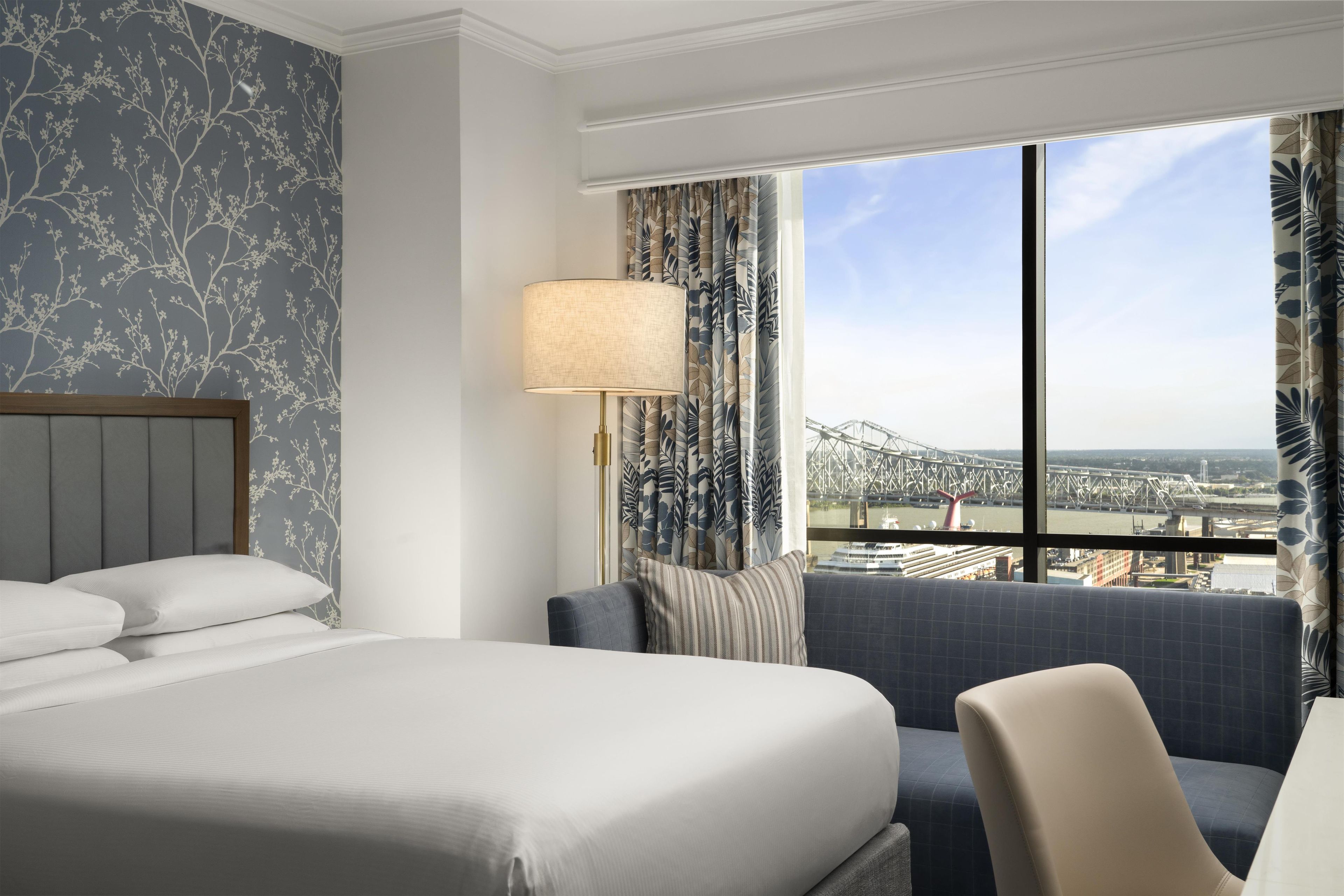 A hotel room with a large bed, a seating area, a lamp, and a view of a bridge.