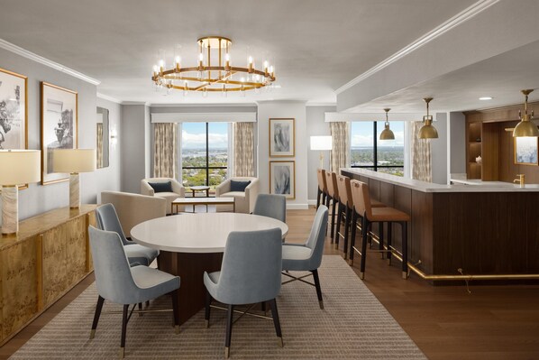 A modern hotel room with a round dining table, bar area, and large windows offering a city view.