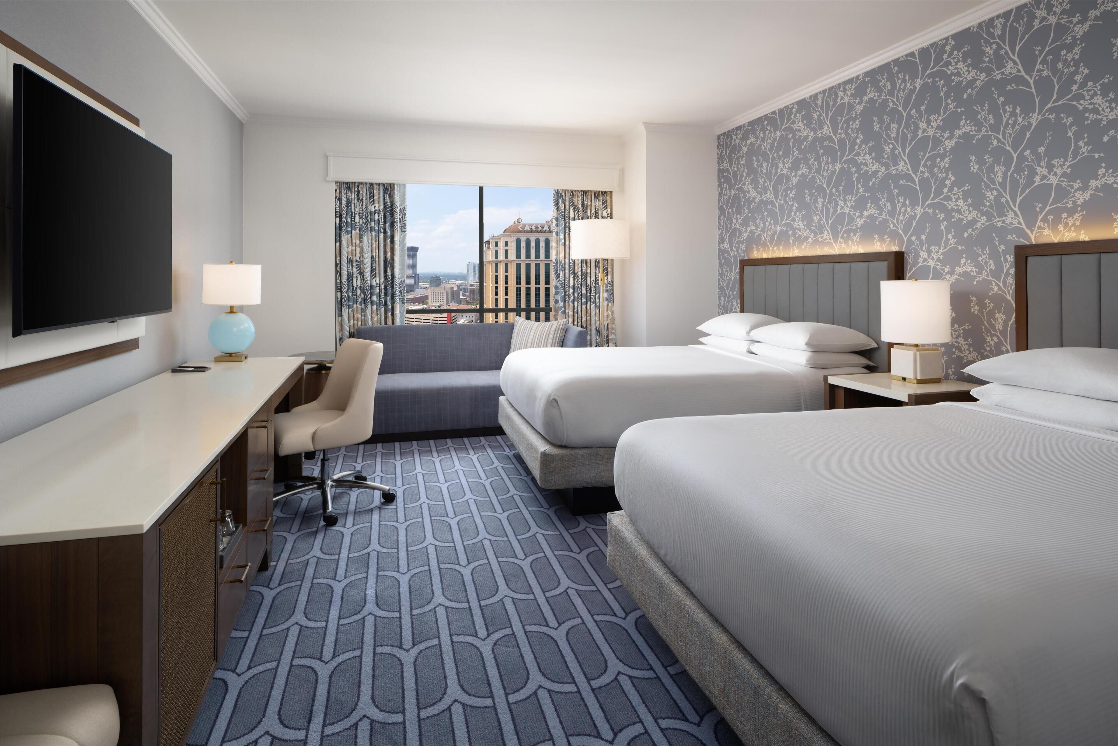 Habitación de hotel con una cama grande, un escritorio, una silla y un televisor. Se ve una ventana con vistas a edificios.