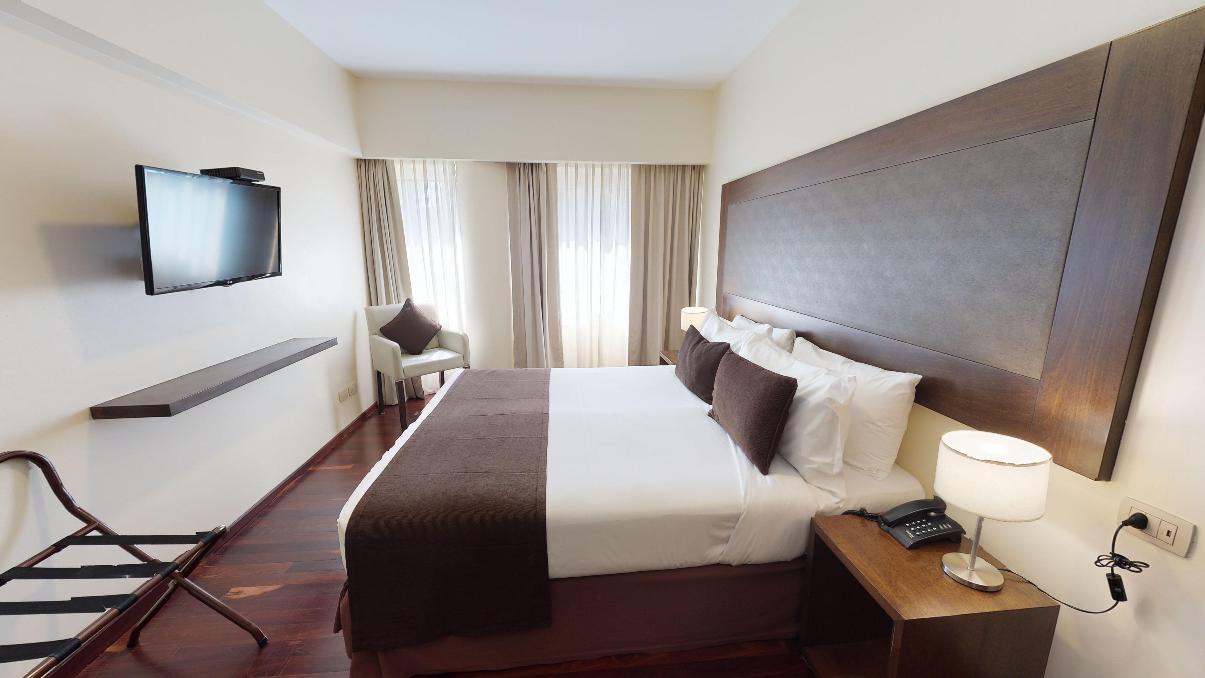 Habitación de hotel con cama, mesita de noche, lámpara, teléfono, televisor, silla y cortinas.