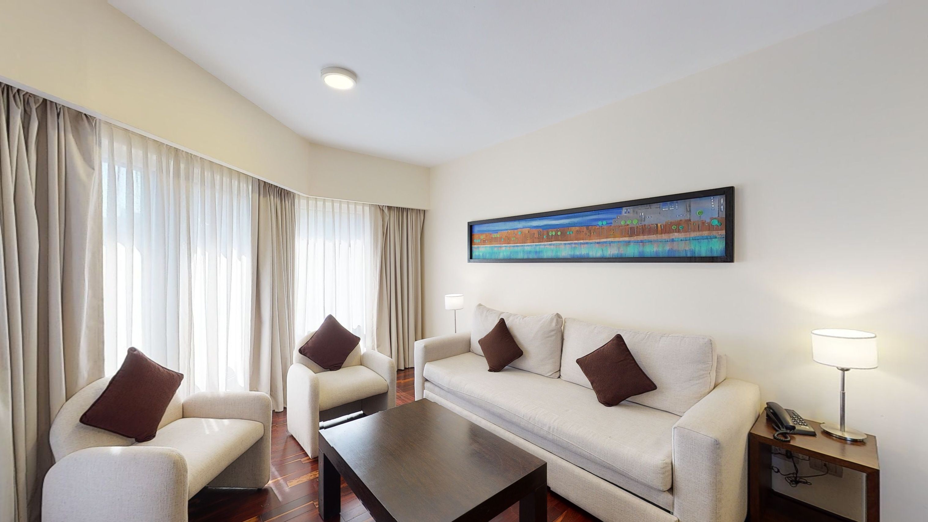 A hotel room with a sofa, armchairs, a coffee table, a painting on the wall, and a bedside lamp.