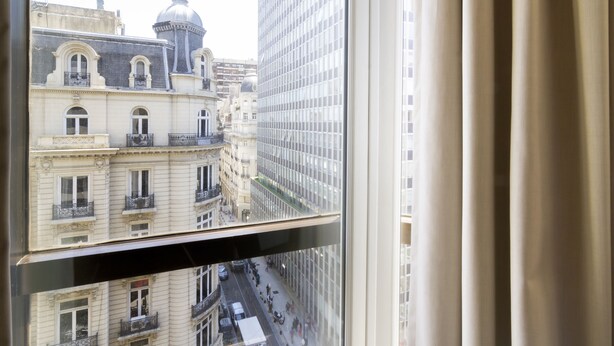 Une vue à travers une fenêtre avec des rideaux, montrant une scène de rue avec des bâtiments et un ciel dégagé.
