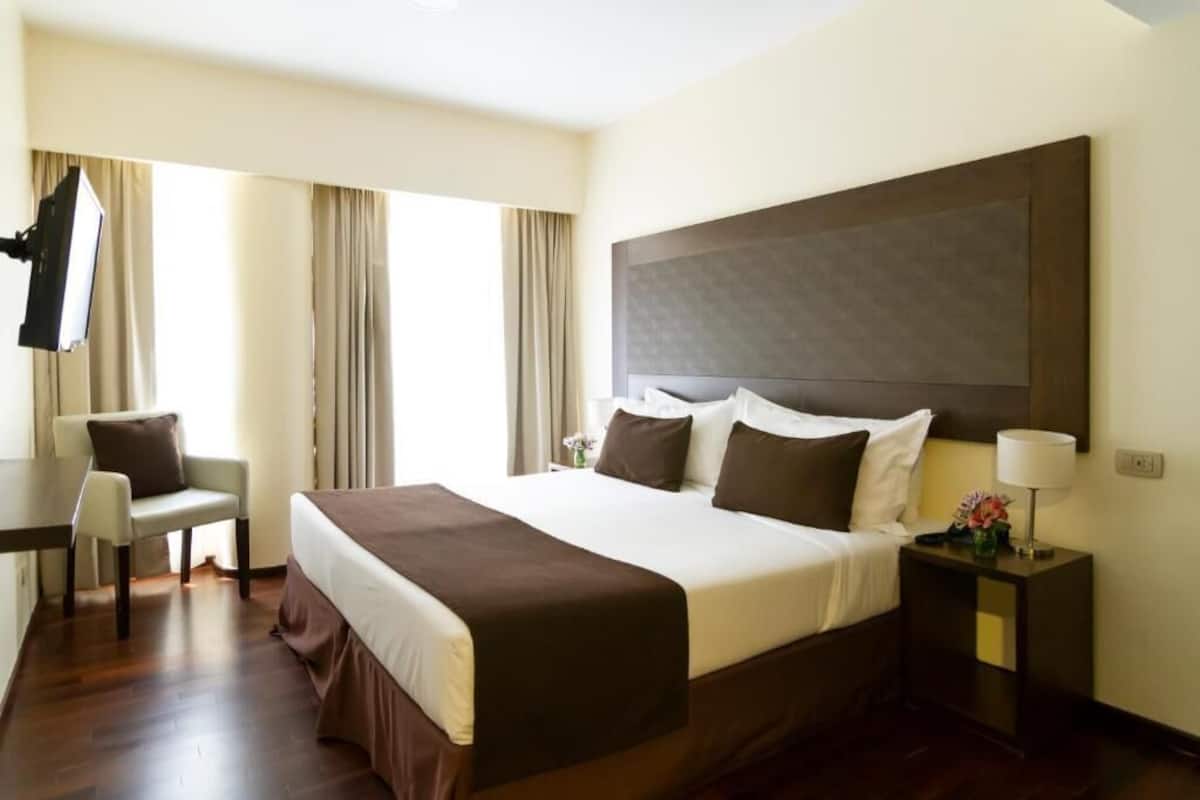 Habitación de hotel con una cama grande, un escritorio, una silla, una mesita de noche y un televisor.